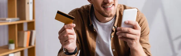 Panoramic shot of businessman using smartphone and credit card in office — Stock Photo