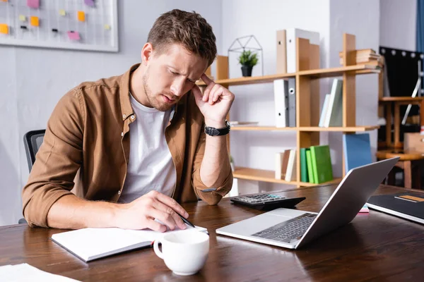 Enfoque selectivo del hombre de negocios pensativo que escribe en el cuaderno al lado de la taza de café, computadora portátil y calculadora en la mesa - foto de stock