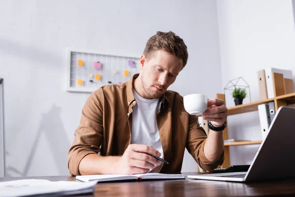 Selective focus of businessman writing on notebook and drinking coffee in office — Stock Photo
