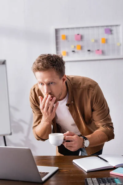 Enfoque selectivo de empresario sorprendido con taza de café mirando a la computadora portátil cerca de papelería en la mesa - foto de stock