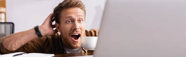 Panoramic shot of excited businessman with coffee cup looking at laptop near notebook in office — Stock Photo