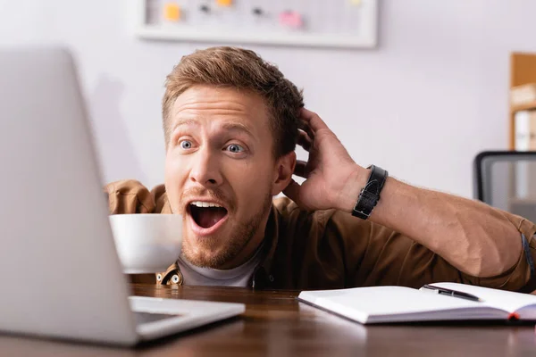 Вибірковий фокус збудженого бізнесмена, який дивиться на ноутбук, тримаючи чашку кави — стокове фото