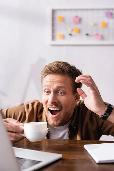 Enfoque selectivo de empresario emocionado mirando portátil cerca de la taza de café y portátil en la mesa - foto de stock