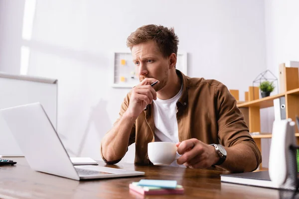 Enfoque selectivo del empresario pensativo que sostiene la taza de café y la pluma mientras trabaja con el ordenador portátil en la oficina - foto de stock