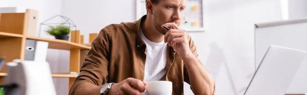 Панорамна концепція продуманого бізнесмена, який тримає чашку кави і ручку біля ноутбука в офісі — стокове фото