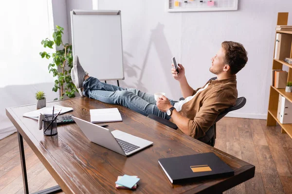 Foco seletivo de empresário segurando xícara de café e usando smartphone perto da mesa de escritório — Fotografia de Stock