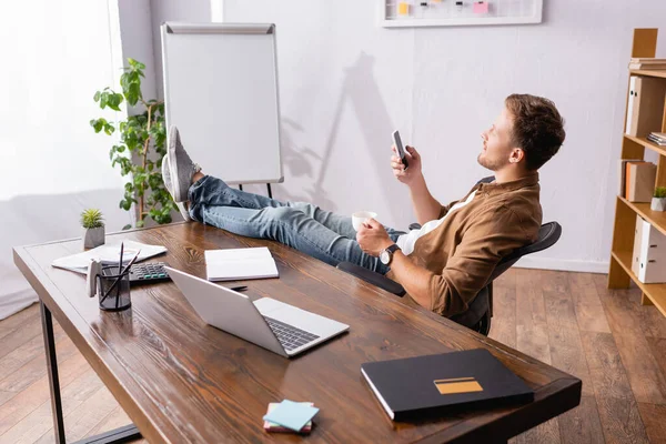 Enfoque selectivo del hombre de negocios sosteniendo la taza de café y utilizando el teléfono inteligente cerca de la mesa de oficina - foto de stock