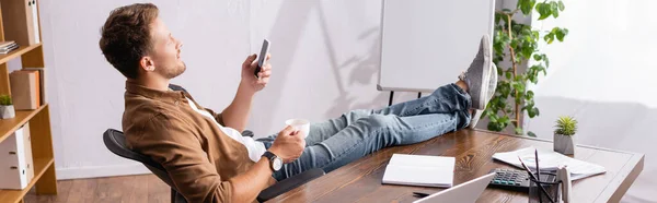 Horizontal crop of businessman using smartphone and holding coffee cup beside stationery on office table — Stock Photo