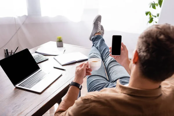 Back view of businessman holding cup of coffee and smartphone with blank screen at office table — Stock Photo