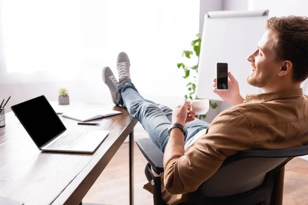 Selective focus of young businessman holding coffee cup and cellphone near laptop on office table — Stock Photo