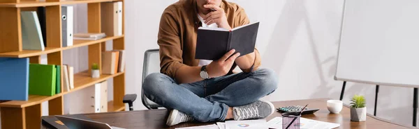 Panoramic crop of businessman holding notebook while sitting on table in office — Stock Photo