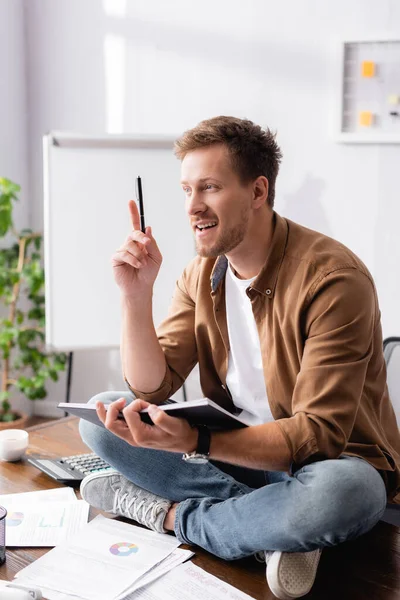 Selective focus of young businessman having idea while holding pen and notebook and sitting on office table — Stock Photo