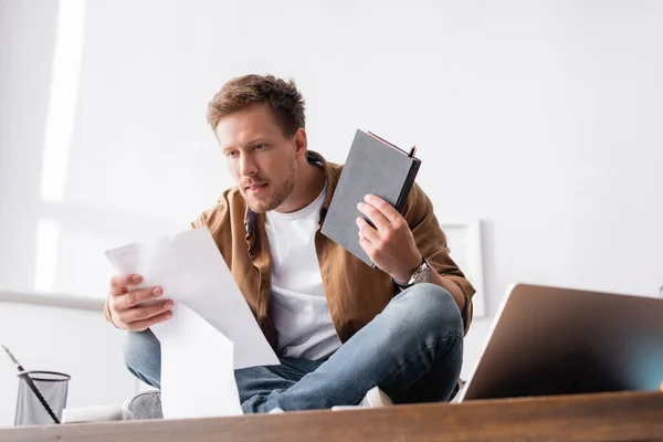 Selektiver Fokus des Geschäftsmannes, der mit Dokumenten und Notizbuch arbeitet, während er mit gekreuzten Beinen auf dem Bürotisch sitzt — Stockfoto