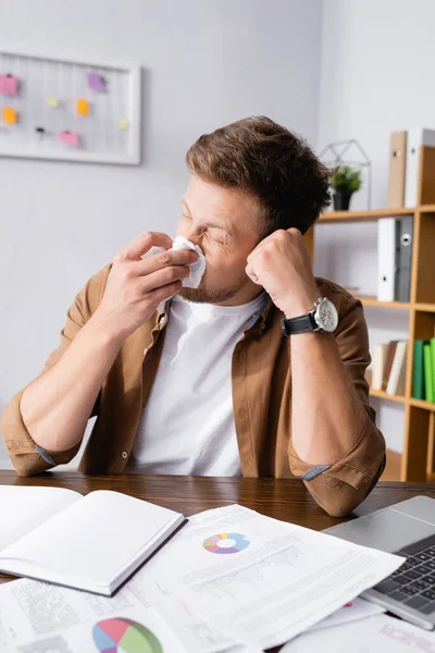 Selective focus of sick businessman holding napkin near nose while working with papers and laptop in office — Stock Photo