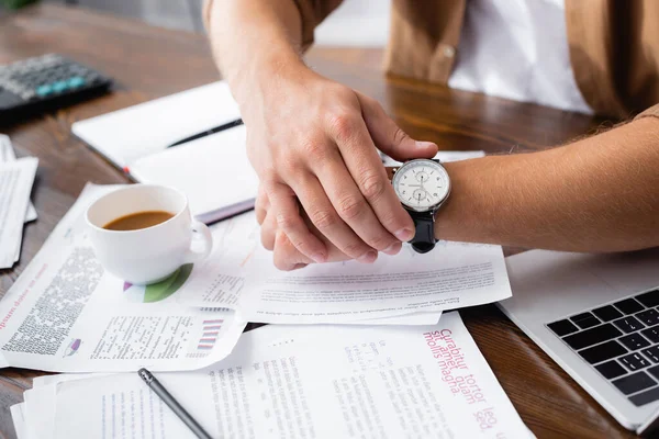 Vue recadrée de jeune homme d'affaires touchant montre-bracelet près des papiers, ordinateur portable et tasse de café sur la table — Photo de stock