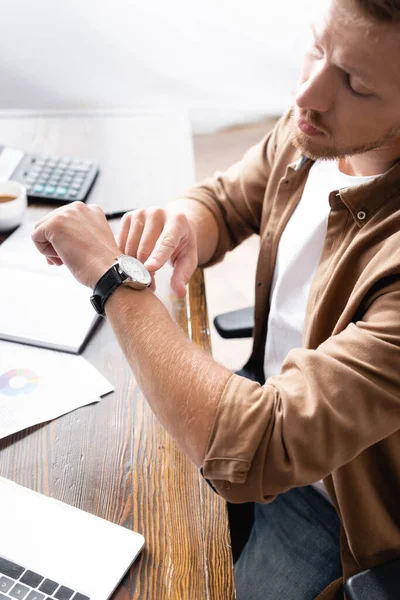 Focus selettivo dell'uomo d'affari che punta l'orologio da polso mentre lavora con laptop e carte — Foto stock