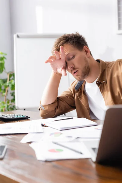 Foco seletivo de empresário exausto com a mão perto do rosto olhando para laptop na mesa de escritório — Fotografia de Stock