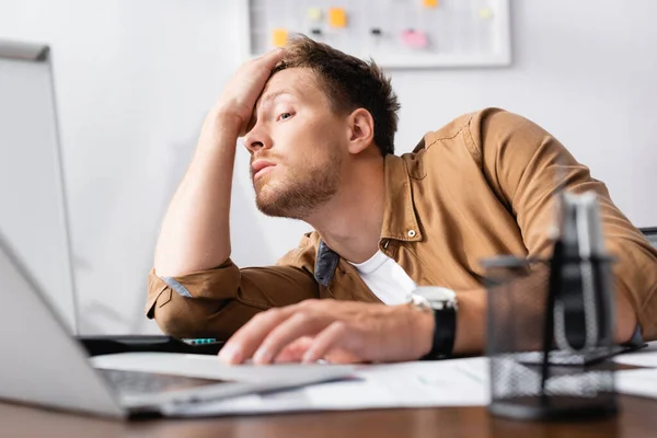 Selective focus of tired businessman with hand near head using laptop while working in office — Stock Photo