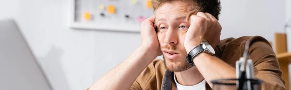 Imagen panorámica del hombre de negocios cansado con las manos cerca de la cabeza mirando a la computadora portátil en la oficina - foto de stock