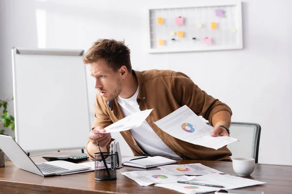 Селективный фокус концентрированного бизнесмена, держащего бумаги и глядящего на ноутбук во время работы в офисе — стоковое фото