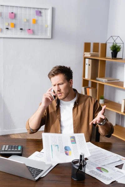 Focus sélectif d'un homme d'affaires sceptique parlant sur smartphone et jetant des papiers au bureau — Photo de stock