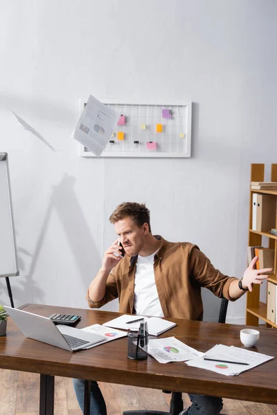 Foco seletivo do empresário irritado jogando papéis enquanto fala no smartphone e trabalha no escritório — Fotografia de Stock