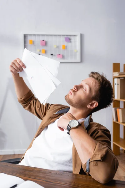 Selective focus of young businessman blowing with documents while feeling hot in office — Stock Photo