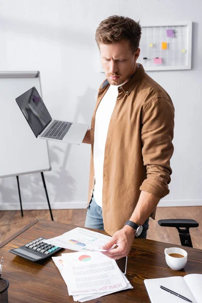Foco seletivo do jovem empresário olhando para papelada e segurando laptop perto de calculadora e café no escritório — Fotografia de Stock