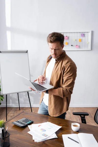 Selective focus of businessman using laptop while standing near papers, calculator and cup of coffee on table — Stock Photo