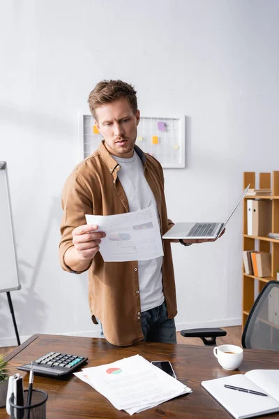 Селективный фокус бизнесмена, смотрящего на документ и держащего ноутбук рядом с ноутбуком и чашкой кофе на столе — стоковое фото