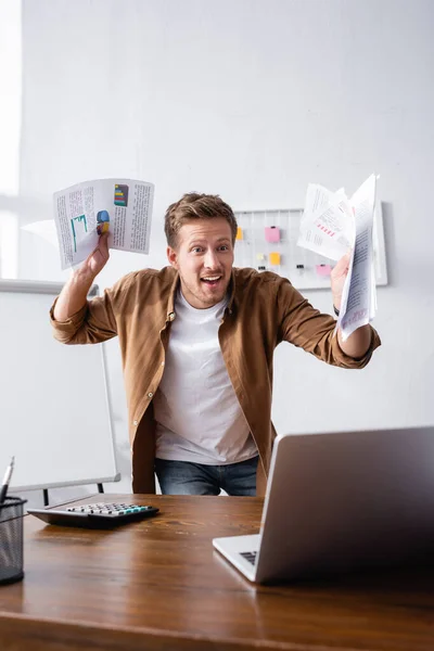 Вибірковий фокус збудженого бізнесмена, який тримає документи біля ноутбука та калькулятора на офісному столі — стокове фото