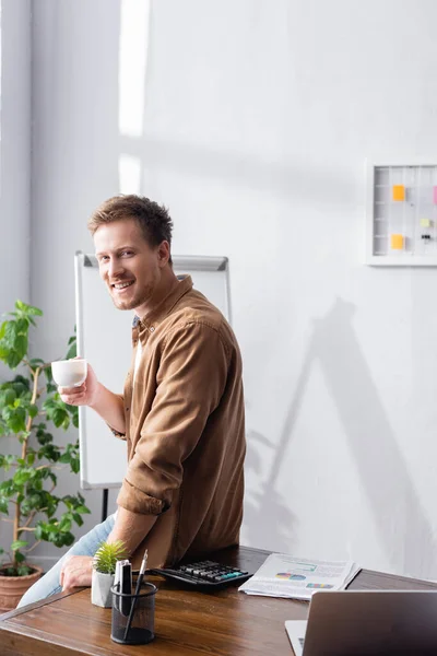 Selektiver Fokus des Geschäftsmannes mit einer Tasse Kaffee, der in die Kamera am Tisch im Büro blickt — Stockfoto