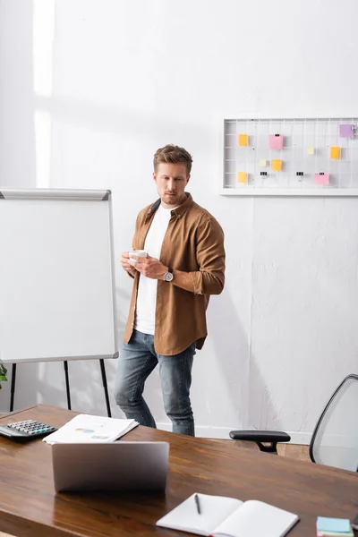 Enfoque selectivo del joven empresario sosteniendo la taza de café cerca de la computadora portátil, papeles y calculadora en la mesa - foto de stock