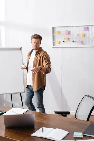 Focus selettivo dell'uomo d'affari che punta con il dito al computer portatile e tiene in mano la tazza di caffè in ufficio — Foto stock