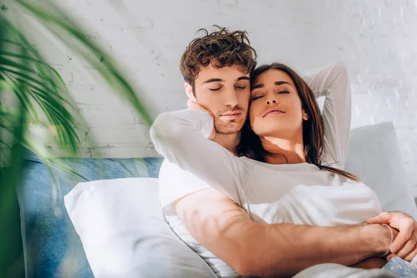 Enfoque selectivo de pareja joven sentada en la cama y abrazándose con los ojos cerrados - foto de stock