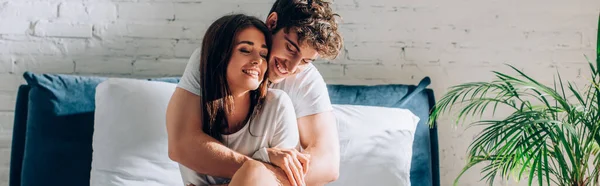 Panoramic shot of young man sitting on bed and embracing girlfriend — Stock Photo