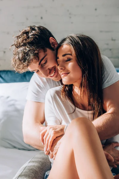 Young man sitting on bed and embracing girlfriend — Stock Photo