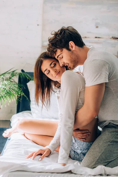 Young man with closed eyes sitting on bed and embracing girlfriend — Stock Photo