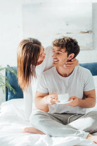 Junger Erwachsener sitzt mit Tasse Kaffee im Bett und schaut Freundin an — Stockfoto