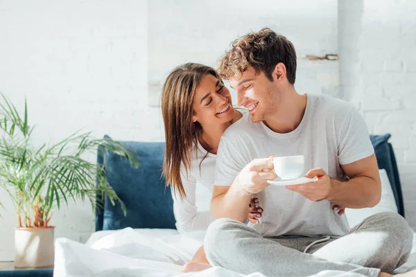 Молодая женщина обнимает и смотрит на парня с чашкой кофе на кровати — стоковое фото
