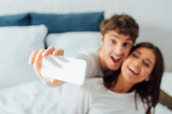 Selektiver Fokus des aufgeregten Paares, das auf dem Bett sitzt und Selfie macht — Stockfoto