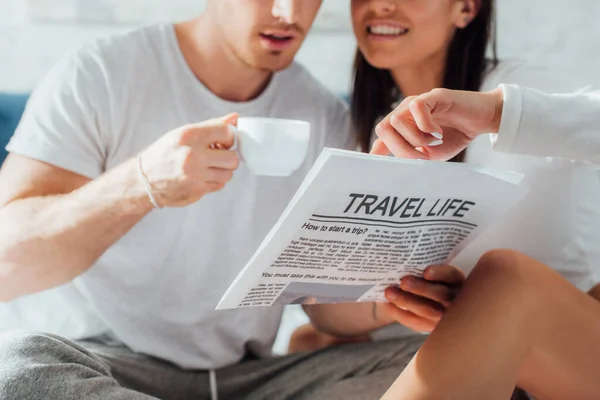 Vista cortada de jovem casal em pijama segurando café e lendo jornal com viagem vida artigo em casa — Fotografia de Stock