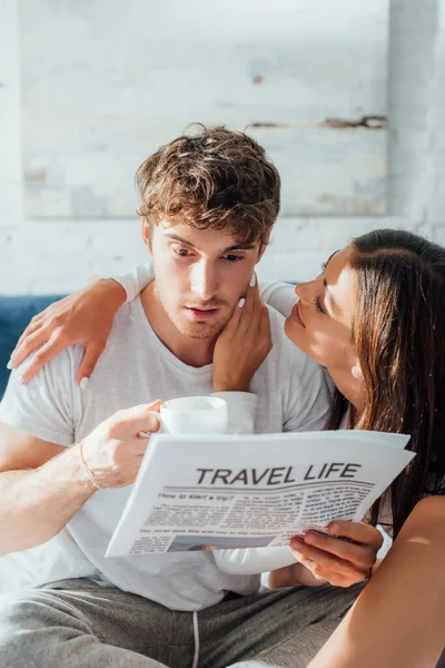 Focus selettivo della donna che abbraccia il fidanzato con tazza di caffè e giornale sul letto — Foto stock