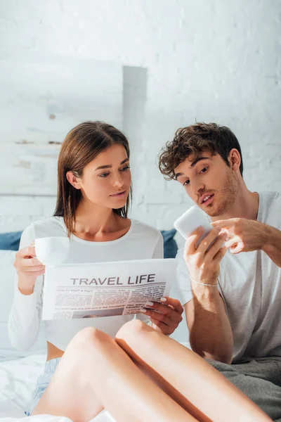 Hombre sosteniendo teléfono inteligente cerca de la novia con taza de café y periódico en el dormitorio - foto de stock