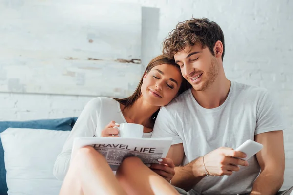 Selective focus of woman holding newspaper and cup of coffee near man with smartphone on bed — Stock Photo