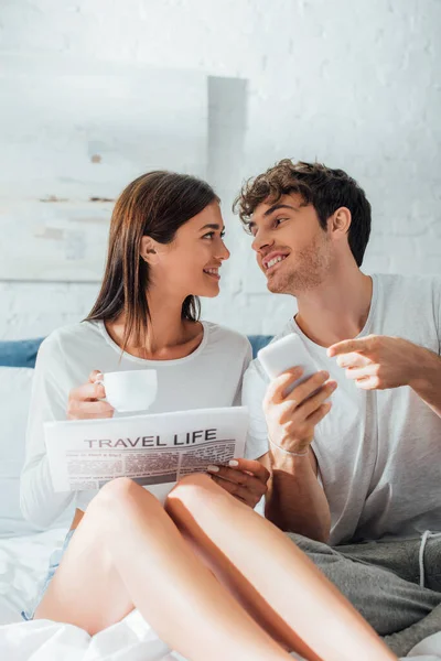 Junger Mann zeigt mit Zeitung und Tasse Kaffee auf Smartphone neben Freundin im Bett — Stockfoto