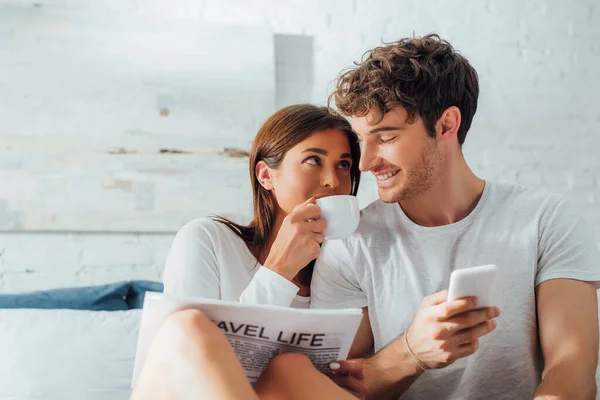 Selective focus of woman with newspaper drinking coffee and looking at boyfriend with smartphone at home — Stock Photo