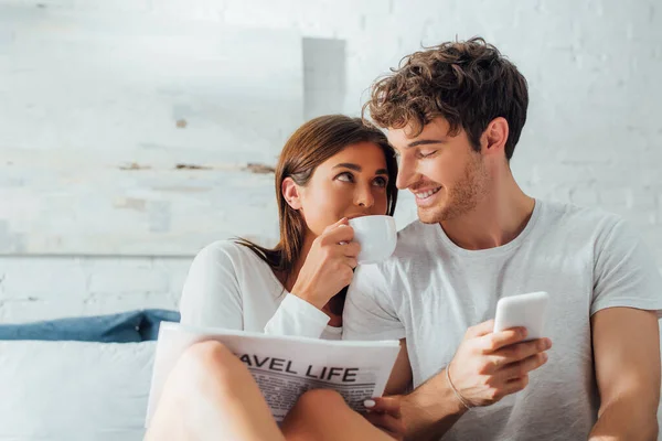 Selektiver Fokus der Frau mit Zeitungskaffee und Blick auf Freund mit Smartphone zu Hause — Stockfoto