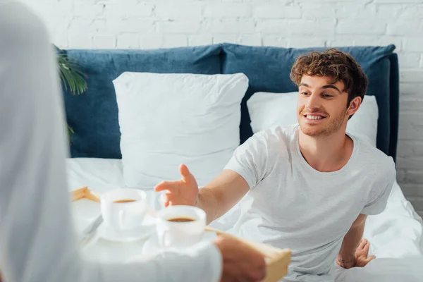 Concentration sélective de l'homme assis sur le lit près de la petite amie avec des tasses à café sur le plateau de petit déjeuner — Photo de stock