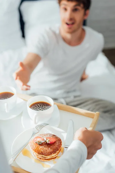 Enfoque selectivo de la mujer sosteniendo panqueques y café en la bandeja del desayuno cerca del hombre en el dormitorio - foto de stock